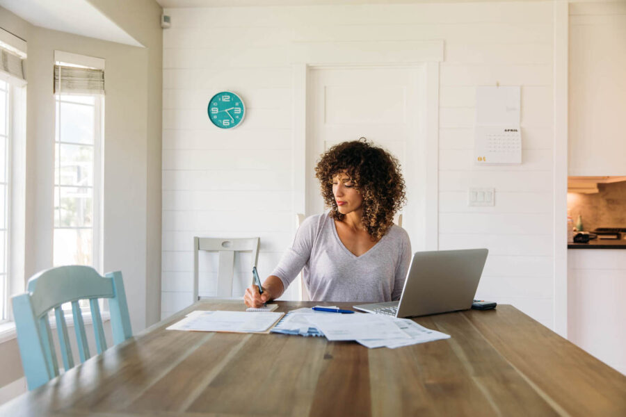 A woman with curly hair writes on a small notepad with documents around it while her laptop sits on the kitchen table.