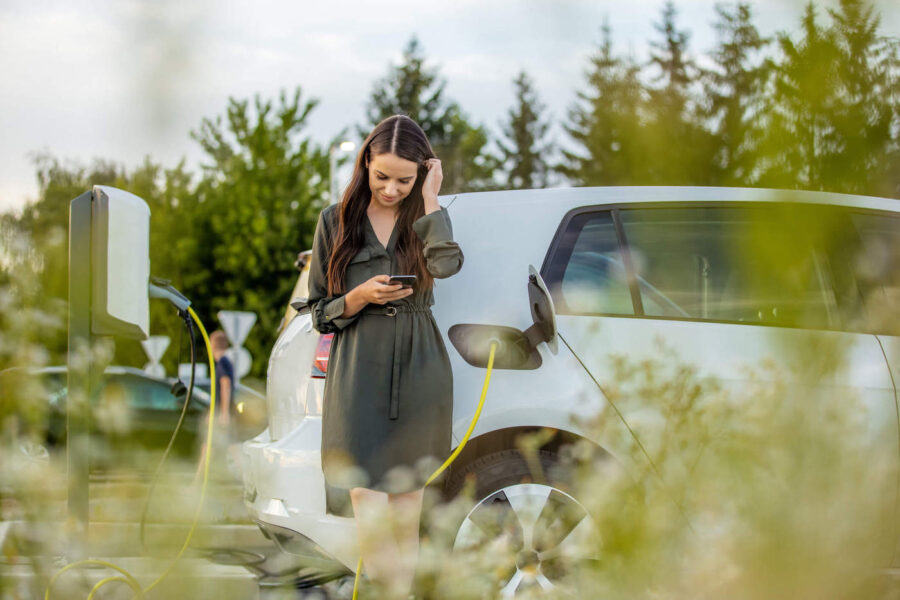 A woman wearing a green dress uses her phone while her electric car charges next to her.
