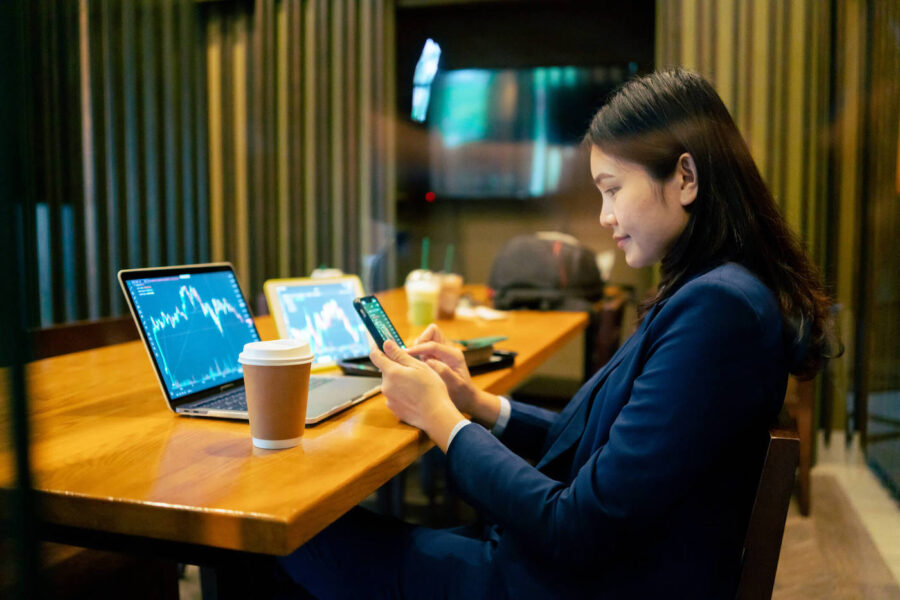 A woman looks at a stock chart on her phone while on the table a laptop and a tablet are both showing stock charts.