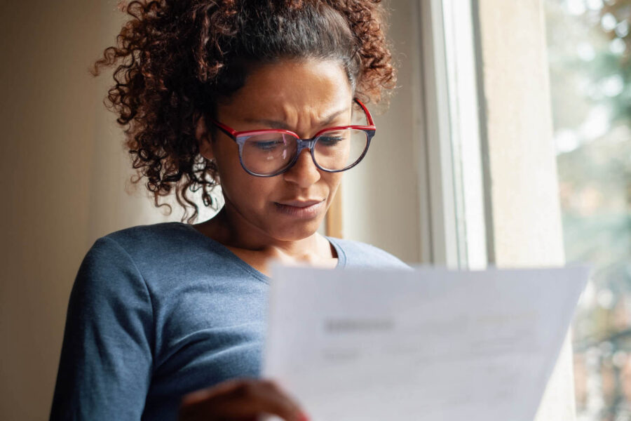 A woman with glasses has a worried look on her face as she look at a document next to her window.