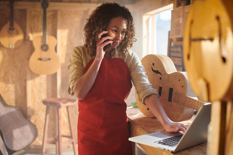 A women with curly hair is wearing a red apron while talking on the phone and typing on her laptop inside her guitar shop.