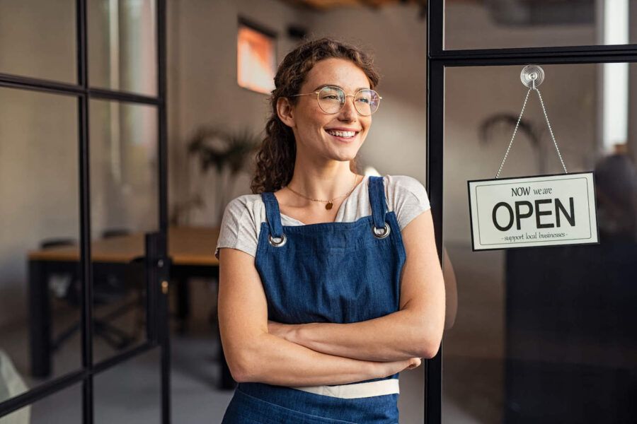 A young business owner women smiles as she wears a blue apron next to an open greeting sign outside of her business.