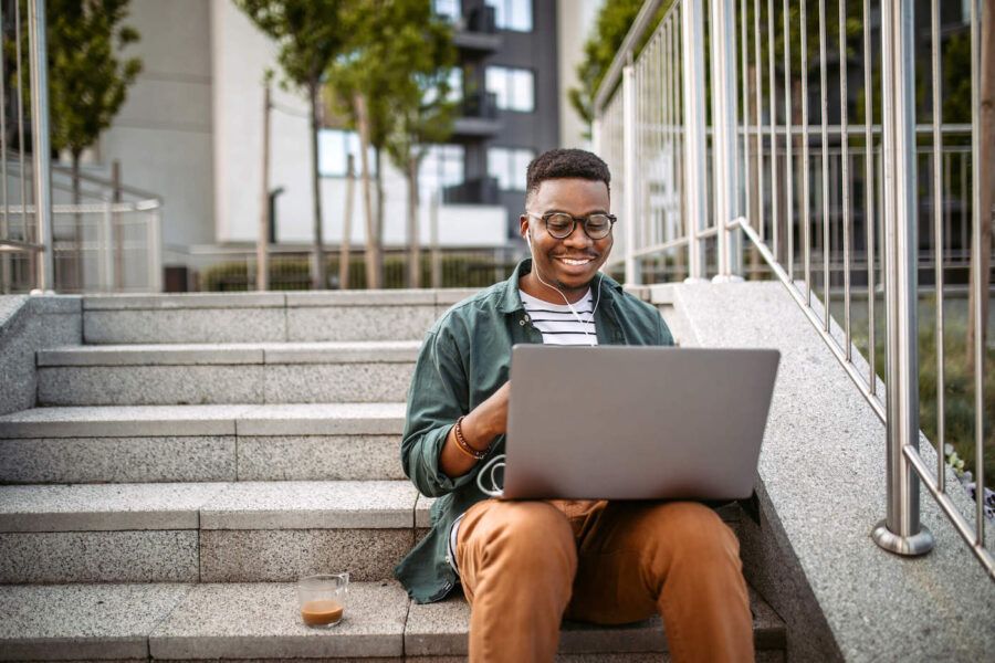 A young man sitting on stairs smiles while using his laptop.