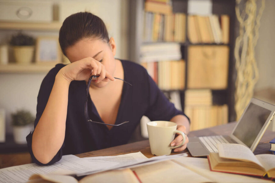 A young woman with her hand on her face has her head down while documents and books are in front of her work desk.