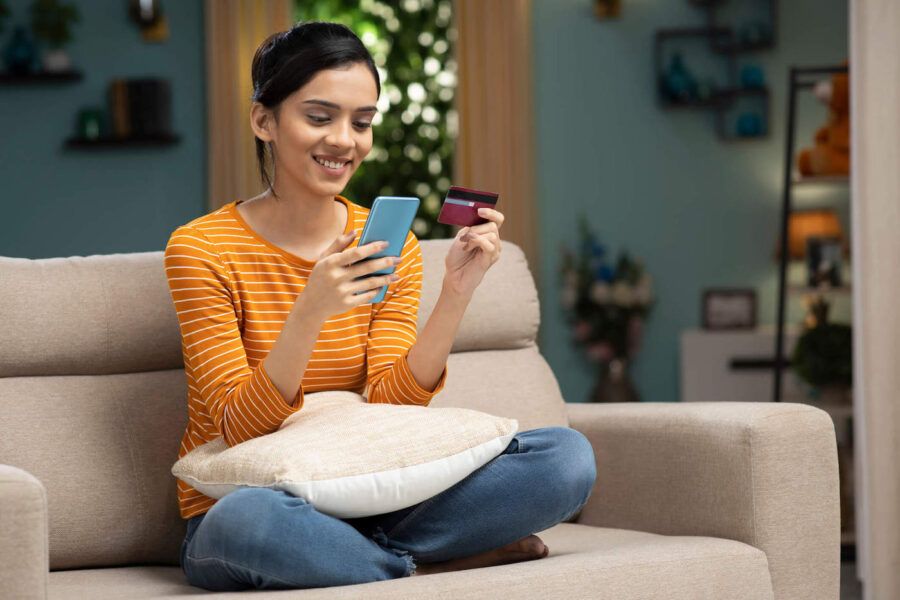 A young woman wearing an orange shirt sits on the couch while smiling at her phone and her other hand holding a credit card.
