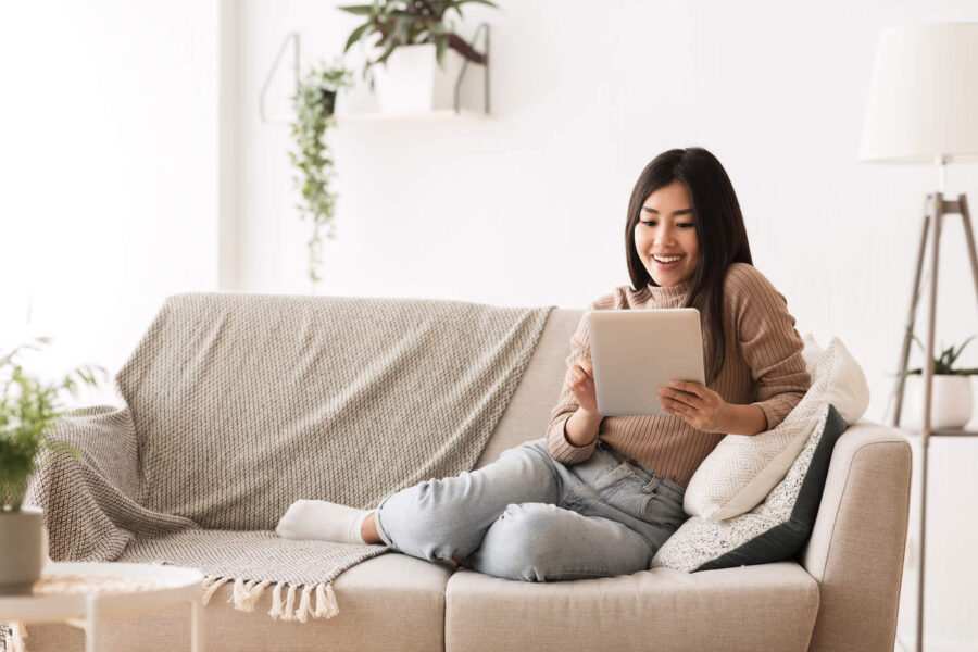 A young woman wearing a beige sweater smiles at her tablet while sitting on the couch.