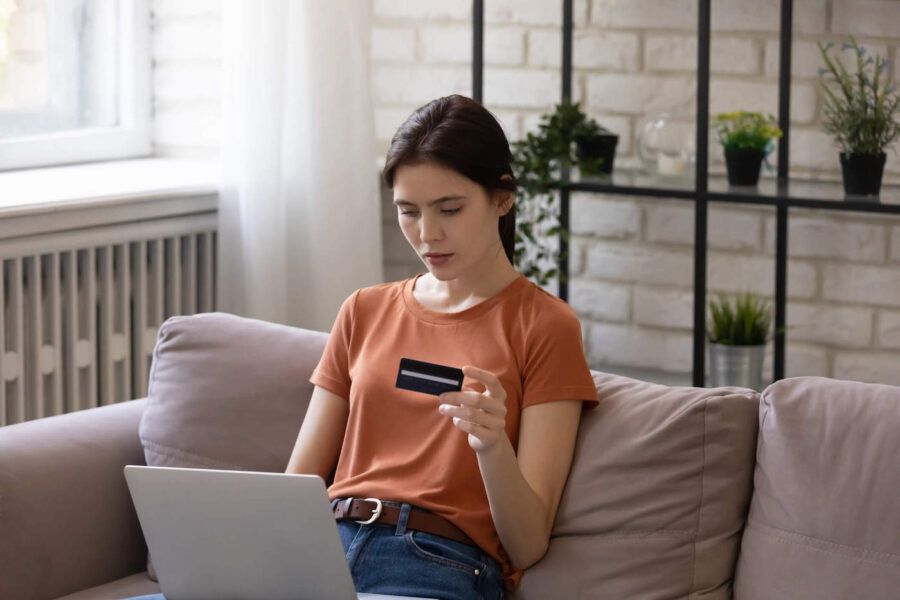 A young woman sitting on the couch uses her laptop while holding her credit card in her hand.