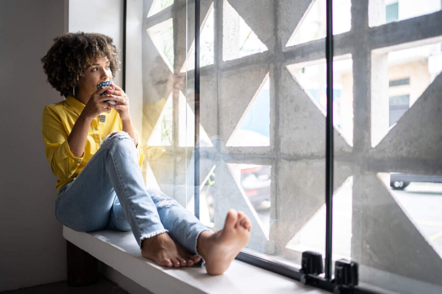 Young woman drinking coffee relaxing on window sill at home.
