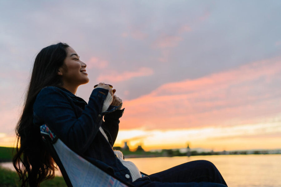 Young woman enjoying hot drink in nature during sunset by lake.
