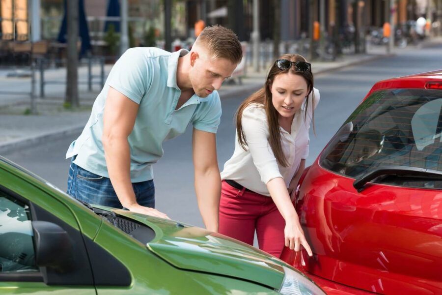 Two people stand on a city street examining damage to the front and rear of two cars after a collision.