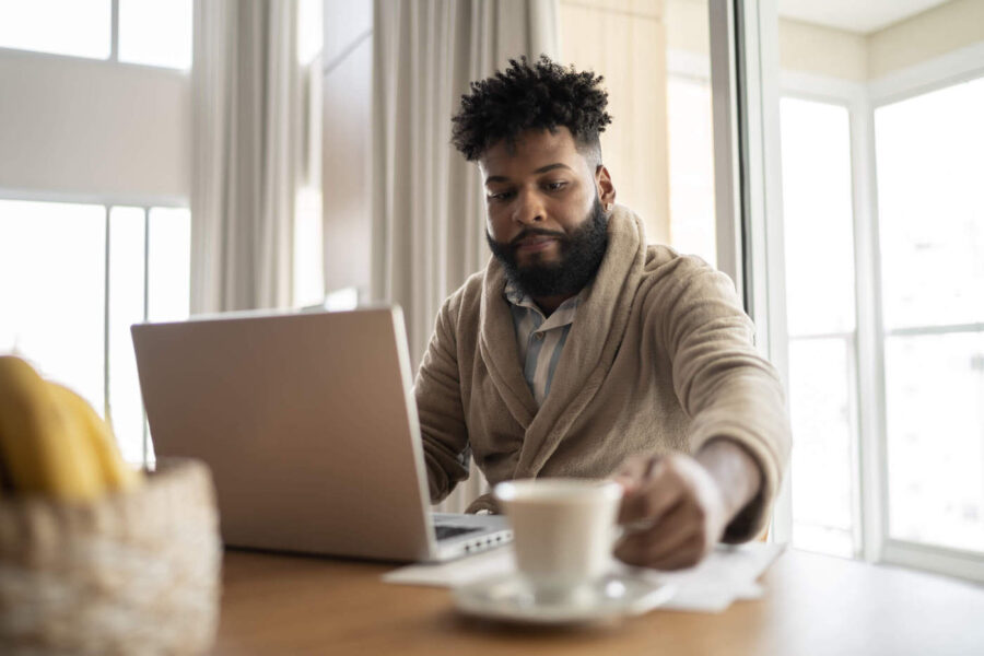 African American man in bathrobe reaching for cup while seated in front of open laptop.