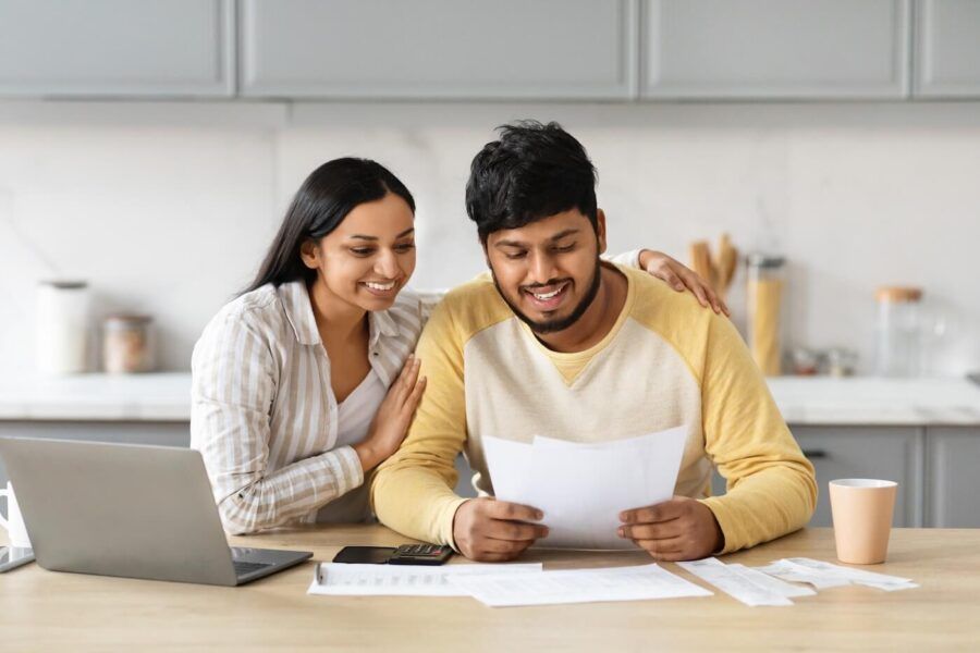 Smiling young couple in a modern kitchen, reviewing printed documents together while seated at a table with an open laptop