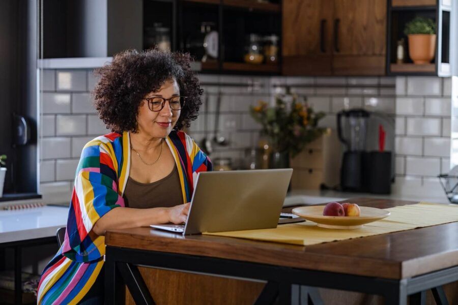 Smiling mature woman in a colorful robe browsing on her laptop in the kitchen.