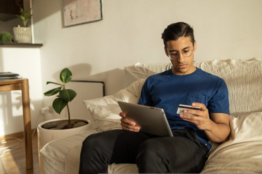 A man with glasses wearing blue shirt in his apartment, sitting on the couch using tablet and looking at the credit card.