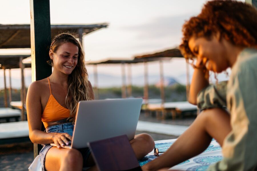 Two women on the beach using laptops during summer time vacation.