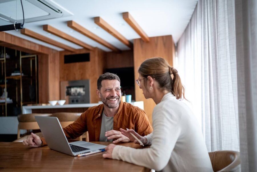 Happy couple sitting at a dining table with an open laptop between them, with the man gesturing while the woman looks at the screen in a modern home interior.
