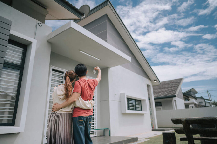 Young couple with their back turned are holding up their new house keys at the front lawn of their new home.