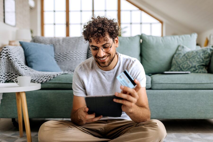 Young man holding a tablet and a credit card while sitting on a floor at home.