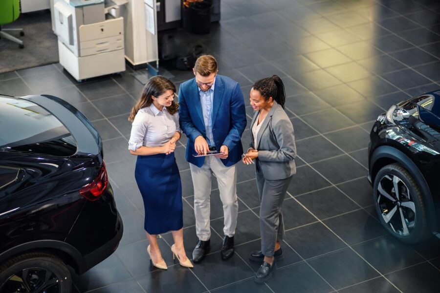 A bird-eye shot of the three agents in the car dealership showroom. The male agent showing something on the tablet to his female colleagues.