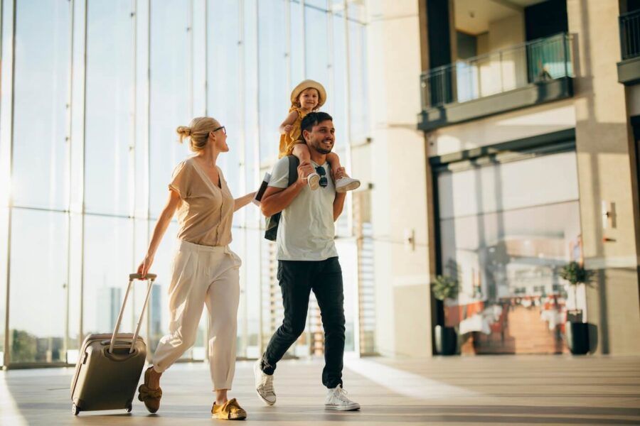 Happy family of three walking through an airport terminal; the young girl is sitting on her father's shoulders, while the mother walks beside them pulling a rolling suitcase.