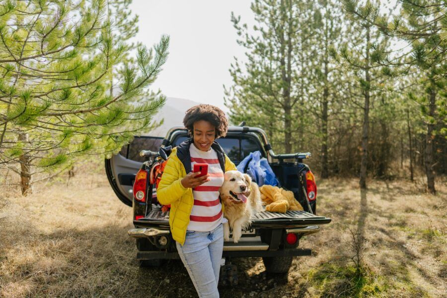 Photo of young smiling woman on a road trip with her retriever dog, riding along on their pick up truck; using her mobile phone.