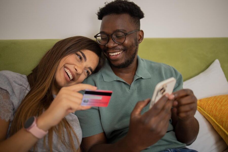 Happy young couple sitting at home, using a smartphone and a credit card to shop online together