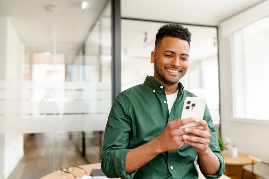 Young man stands with a smartphone in hands, beaming with a smile as he looks at the screen, investing online.