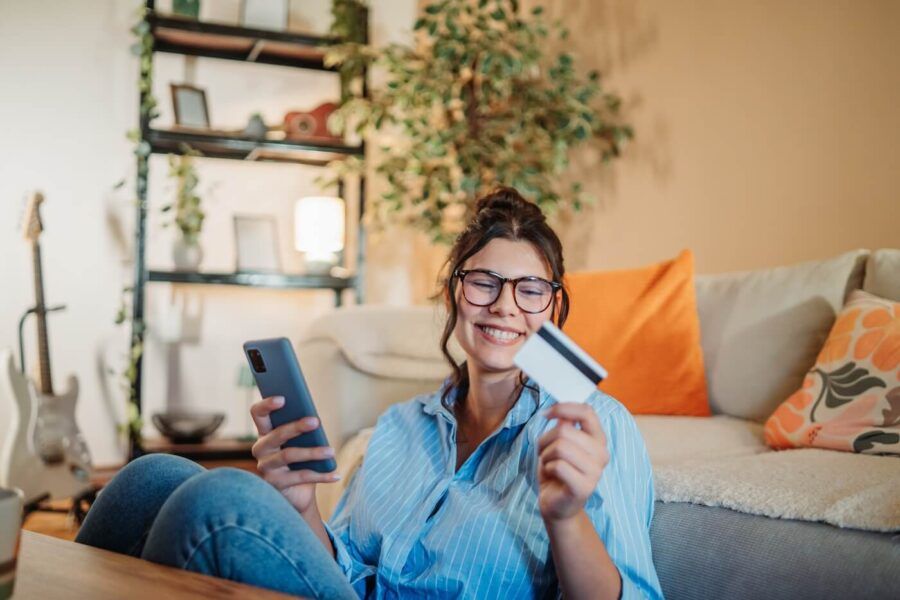 Smiling young woman sitting on the floor next to a sofa in a cozy living room, using her credit card and a smartphone