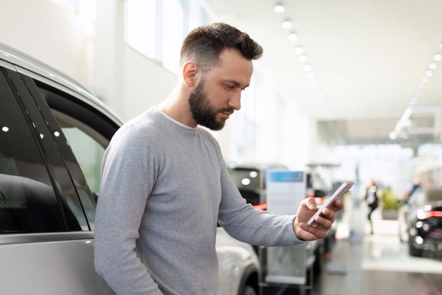 Young man standing next to a white car in the dealership and checking his smartphone