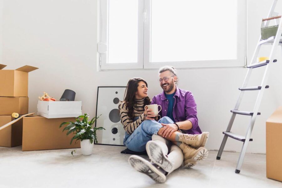 Happy young couple sitting on the floor of their new living room surrounded by potted plans, paintings, and boxes