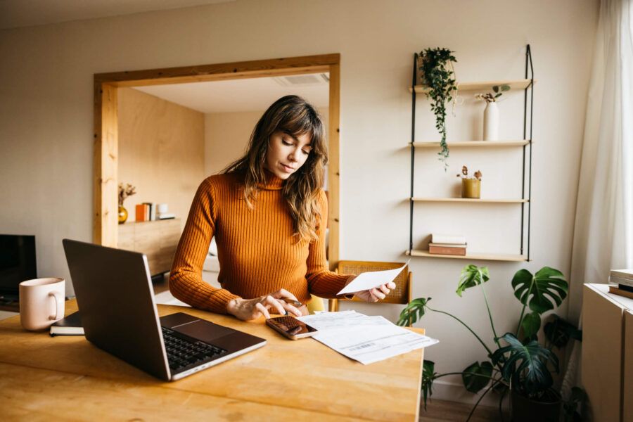 Focused woman sits at a wooden table using a calculator while reviewing printed documents, with a smartphone and laptop nearby in a bright home workspace.
