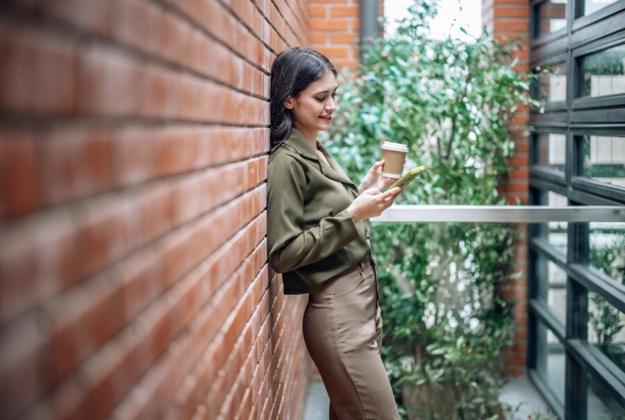 A woman checking her credit report on her phone, holding a coffee and leaning up against a brick wall.