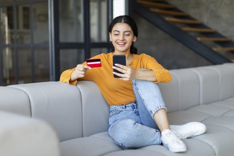 Relaxed young woman using credit card and mobile phone while sitting on the couch.
