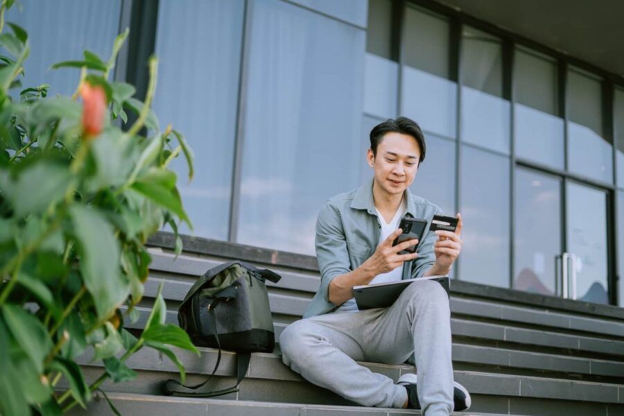 Smiling young man sitting on outdoor stairs with a tablet on his lap, using his mobile phone and holding a credit card, with a backpack placed beside him