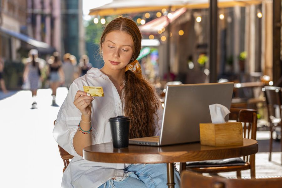 Girl holding credit card and using laptop computer outdoors. Young female tourist sitting at table in a cafe in the city