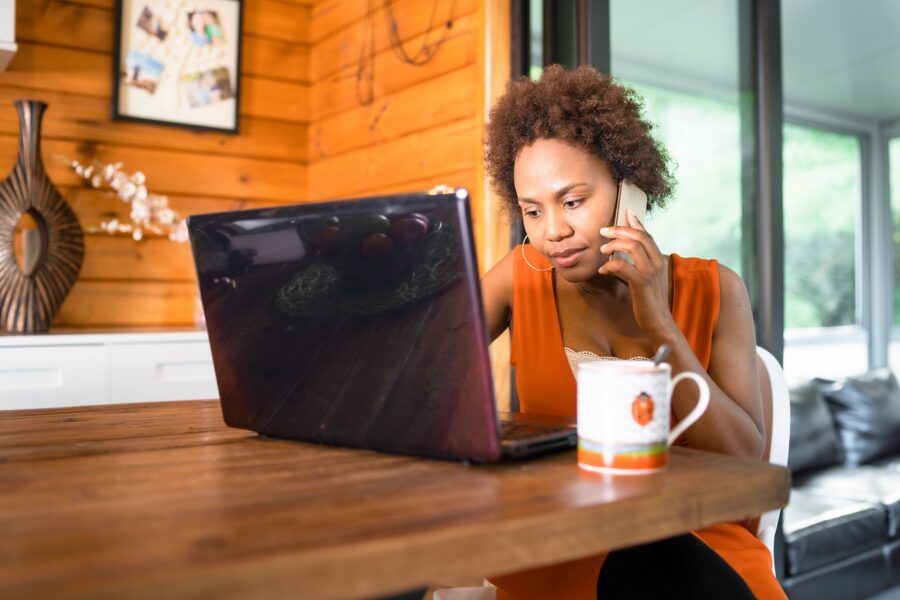 Woman working from home on her mortgage payments. She is sitting at a wood table with a cup of coffee beside her, typing on a laptop and talking on the phone.