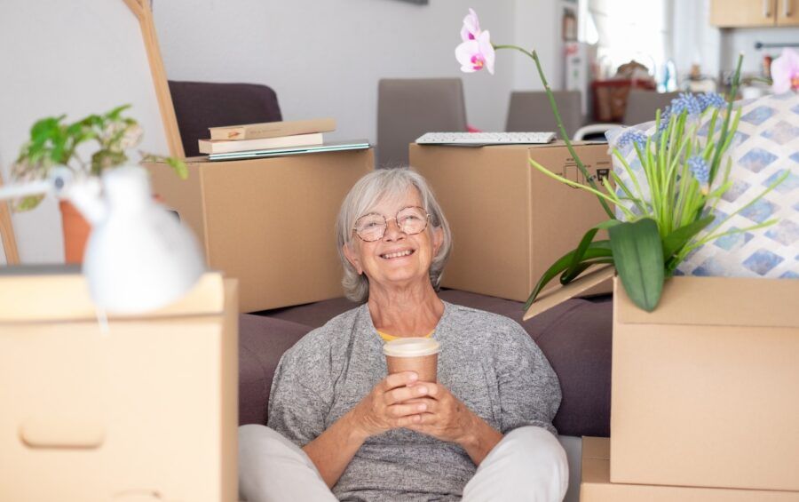 Happy senior woman involved in moving house sitting among cardboard boxes takes a break with a coffee.