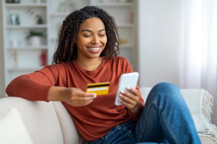 Smiling young woman sitting on a white couch and using a credit card on her smartphone