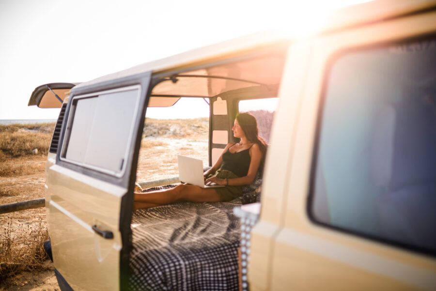 A woman buying car insurance online using her laptop while sitting in the back of her camper van parked in a field at sunset.