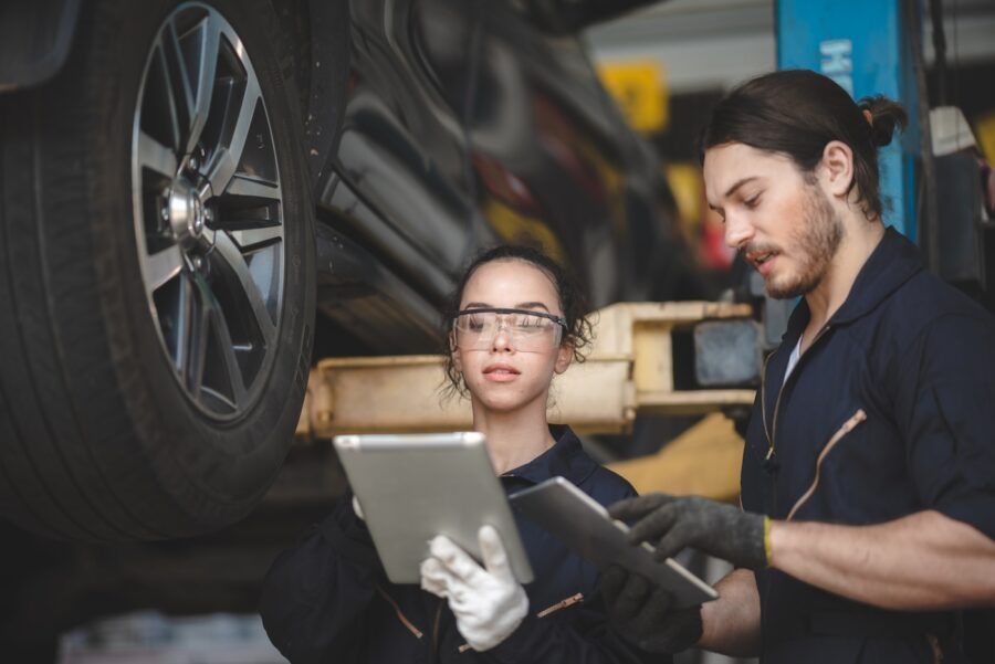 Young male and female car mechanic wearing uniform with safety eye glasses and hand gloves working on digital tablet while diagnosing problem in car in garage and discussing solutions.