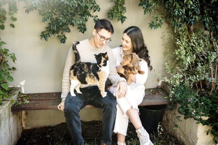 Happy young couple with their pet dog and cat sitting on wooden bench in backyard of their home.