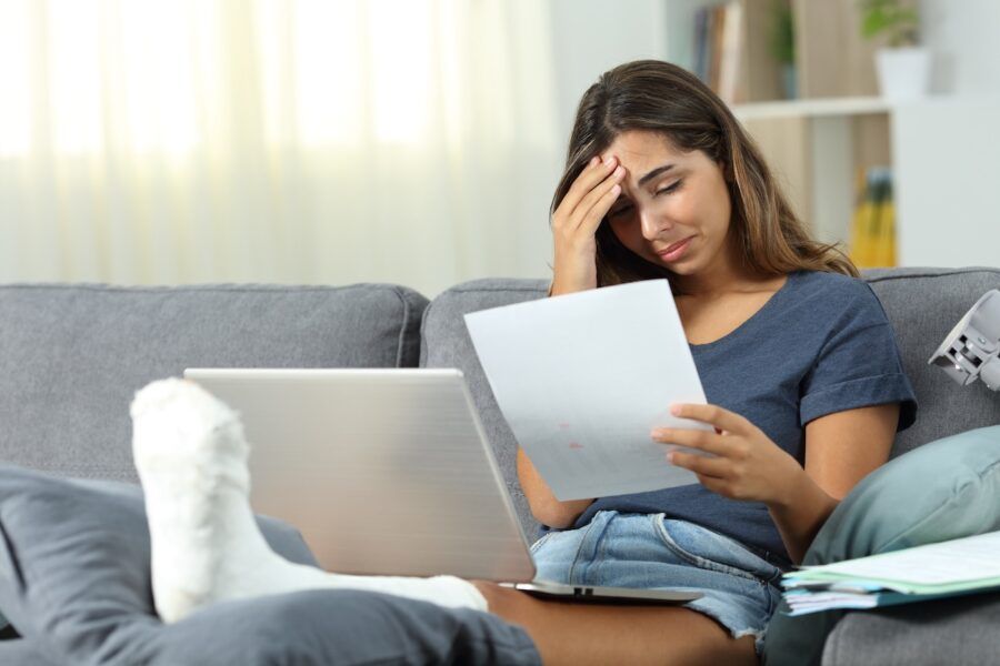 Worried woman with a broken leg reviewing her medical debt on the couch.