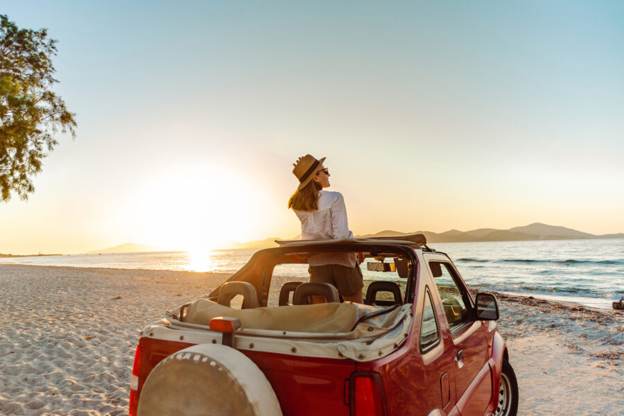 Cheerful young woman in an open-top car parked on the beach. She is enjoying the sunset and looking at the sea