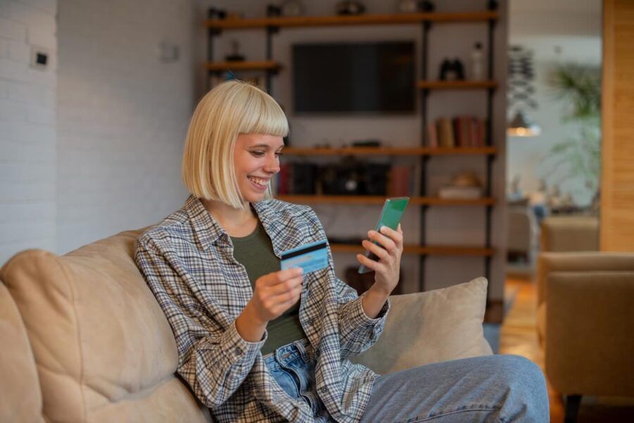 Smiling young woman sitting on a sofa and using her credit card in the mobile app