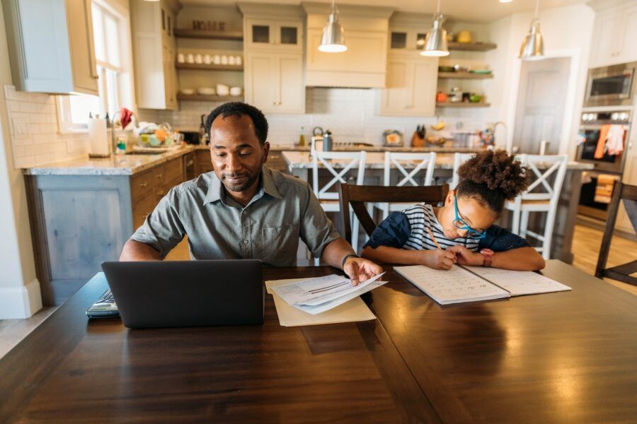 A middle-aged man sits at his dining room table with his young daughter doing his home finances.