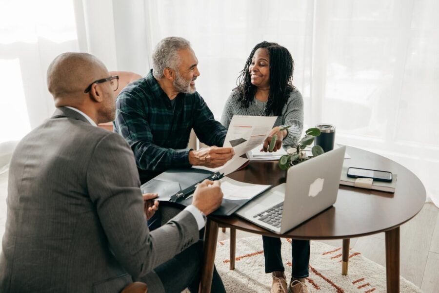 Two clients seated at a round table reviewing documents with a financial professional, with a laptop, paperwork, folders, and a smartphone visible during a consultation in a bright home office setting.
