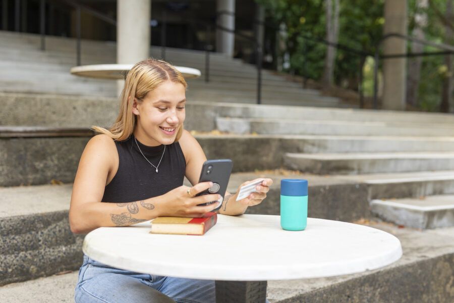 Young college girl using her student credit card on her smartphone while sitting at a table near amphitheater steps.