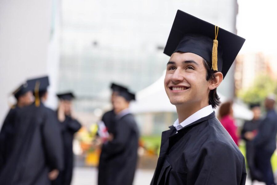 Smiling young man wearing a graduation cap and gown at an outdoor commencement ceremony, standing among other graduates with a campus building and attendees visible in the background.