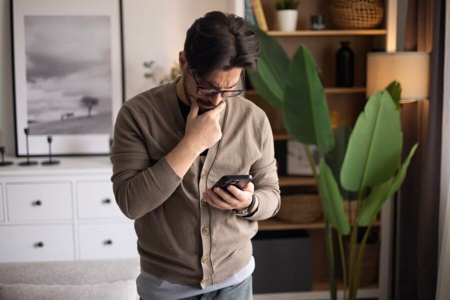 Concerned man standing in a living room and looking at a smartphone while resting a hand on the chin. Bookshelves, framed wall art, indoor plants, and a sofa are visible in the background.