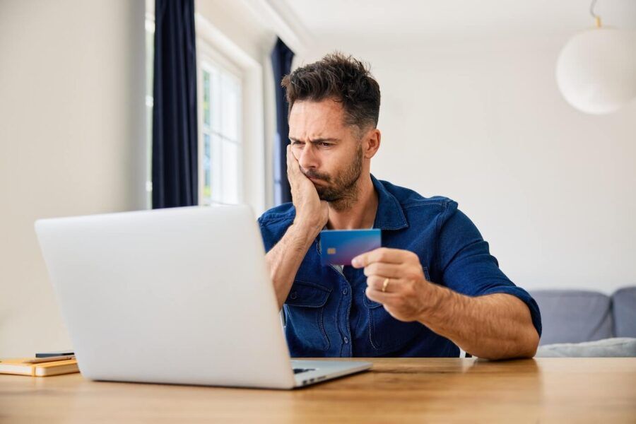 Concerned man seated at a table holding a credit card while looking at a laptop screen in a home setting. A notebook and pen sit beside the computer, with large windows and a sofa visible in the background.